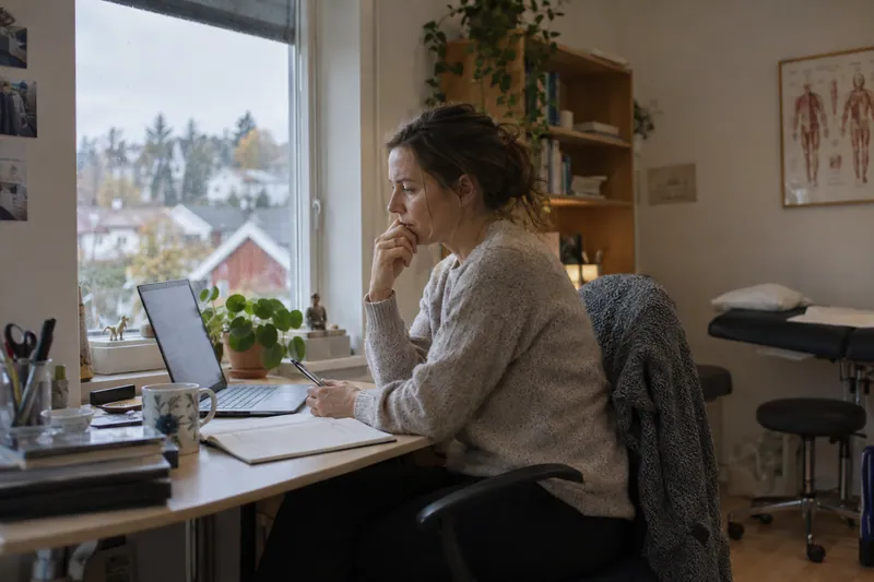 Behandler med notatbok og bærbar PC ved vinduet i et lite kontor.